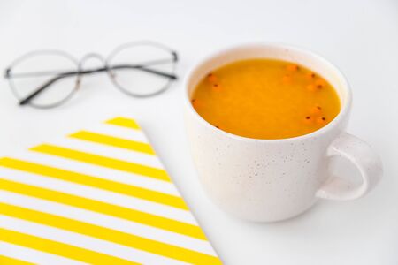 A cup of hot sea buckthorn tea with fruit juice with a yellow notebook and glasses on a gray table. Autumn seasonal layout workplaceの写真素材