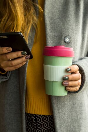 Bamboo eco cup in the hands of a woman in a gray coat and a yellow autumn sweater with a smartphone in her handsの写真素材