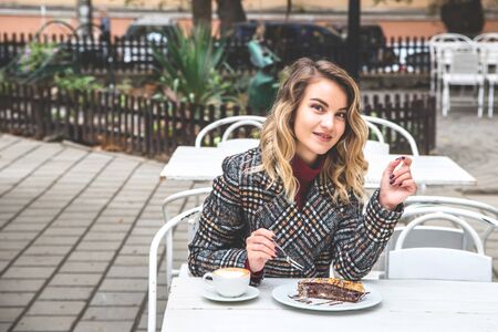 Beautiful young Caucasian having a rest with cappuccino and a piece of cake on the street table of a small cafe on an autumn day.の写真素材