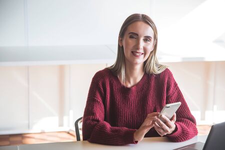 Modern business woman in co working interior working at her smartphone and laptop and smiling at the camera. Freelancer remote work concept.の写真素材