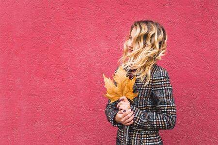 Caucasian blonde girl with curly hair holds a wedge leaf in her hands. Melancholy autumn mood. On a bright red background. Copy space.の写真素材
