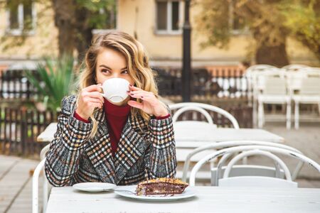 Beautiful young Caucasian having drinks cappuccino and covers his face with a cup sitting outside the cafe on an autumn day.の写真素材