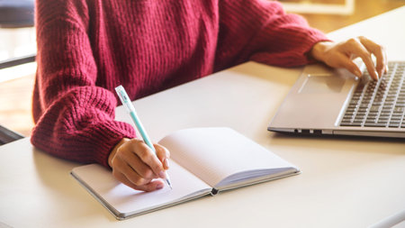 Female hand writes in a white notebook next to a laptop at her workplace as a blogger, journalist or...