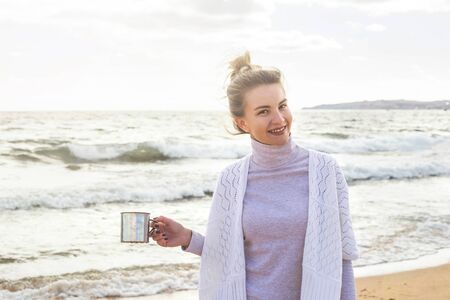 Caucasian girl in a white boho jacket with a bunch on her head and a metal cup in her hand by the sea on a windy day. Traveling in nature.の写真素材