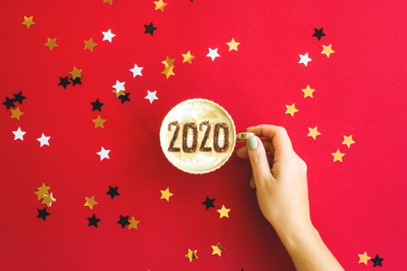 New Year 2020 is written on a cup of coffee with milk in female hands on red background with...