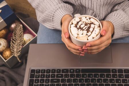 Girl holds a mug of cocoa with marshmallows next to a laptop sitting on the floor in a Christmas interior. Freelancer work remote from home.の写真素材
