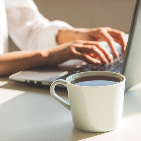 A modern freelancer workplace with a laptop and a cup of tea on a white table in the shadows in a light interior.の写真素材