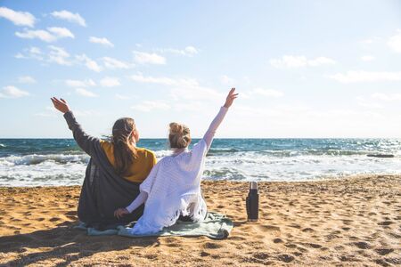 Two young women are sitting on the sand by the sea with their backs to the camera and enjoying a sunny cool day.の写真素材