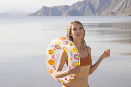 Beautiful millennial girl with a circle for swimming at the sea smiles happily and looks at the camera while standing by the sea.の写真素材
