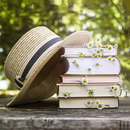 A stack of books on an old wooden table next to a straw hat of a canoe and a bouquet of wild flowers.の写真素材