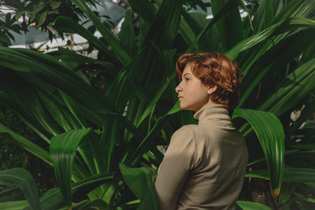 A beautiful plus size girl with red hair among green branches of tropical plants