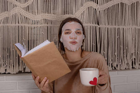 A woman in a facial mask is reading a book and drinking tea on the bed at homeの写真素材