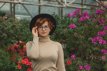 A beautiful plus size girl in eyeglasses and hat smiling among the green plants and flowers.の写真素材