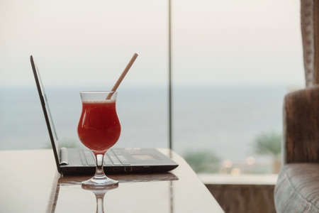 A strawberry cocktail next to a laptop in a bar overlooking the sea. Remote work on vacation concept.の写真素材