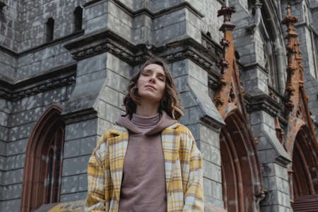 Happy millenial woman in a yellow coat in European city near historic building.の写真素材