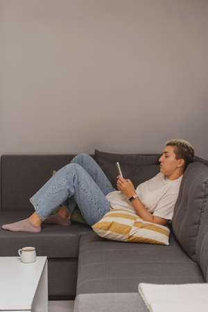 A woman lies on a sofa with a smartphone in a minimalistic home interior.の写真素材