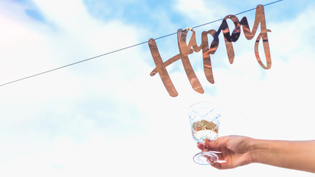 Golden word happy hangs on garland against background of blue sky with clouds next to woman hand with glass of wine.の写真素材