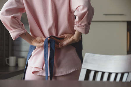 Woman in pink suit ties blue apron on her back to start cooking in her home kitchen. Faceless image.の写真素材