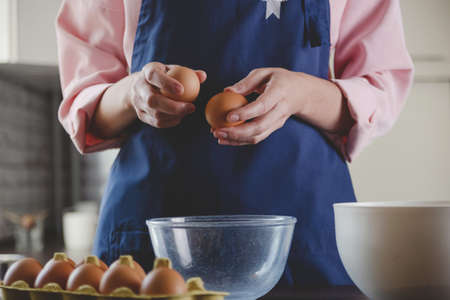 Woman baker in a blue apron breaks eggs for baking. Home cozy cooking aesthetics.の写真素材