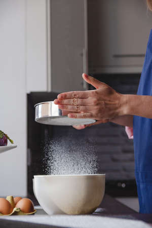 Woman baker in blue apron sifts flour in home kitchen to prepare dinner. Flying flour aesthetics.の写真素材