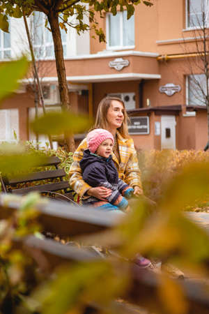 Mom and daughter are sitting on city bench during autumn walk. Fall colorsの写真素材
