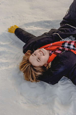 Happy young blond woman lies in snow in black coat, orange scarf and yellow gloves. Winter season. Close upの写真素材