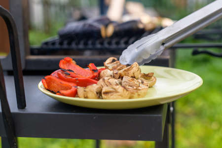 A man spreads grilled bell peppers and mushrooms on a round ceramic plate with kitchen tongsの写真素材