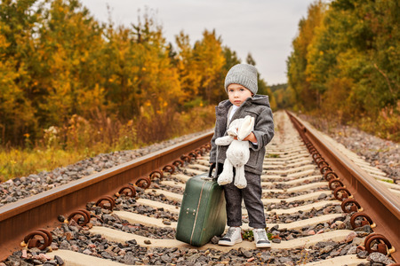 cute sad boy in retro clothes with a vintage green suitcase standing on a rails in the forestの写真素材
