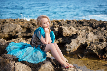pensive girl in azure dress with a tail like a bird sitting on a rocky seacoast of the ocean seaの写真素材