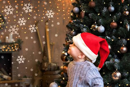 Little smiling boy toddler in a grey knitted sweater and hat of Santa Claus with a background of Christmas tree and snowflakesの写真素材