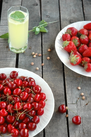 strawberries, sweet cherries and cherry-stones, glass of lemonade with mint leaf and branch  twig  on wooden backgroundの写真素材