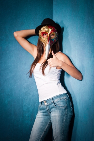 teen girl wearing Venetian mask,black hat and blue jeans stand in corner of room with thumb upの写真素材