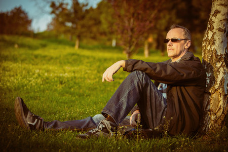 middle-aged man enjoys watching the sunset sit by tree in parkの写真素材