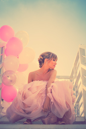 young beautiful woman in elegant wedding dress hold balloons sitting on  white stairs against blue sky, retro colorsの写真素材