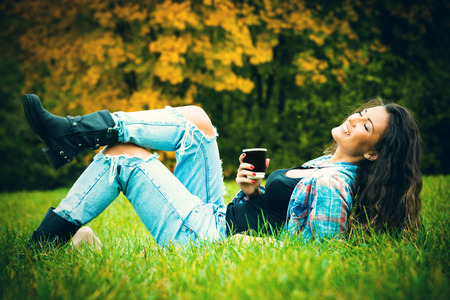smiling young woman enjoy in coffee break in nature, lie on grass in park, wearing blue torn jeans and tartan shirt, full body shotの写真素材