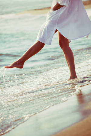 barefoot woman enjoy in sea water in white long shirt, lower body, selective focus, side viewの写真素材
