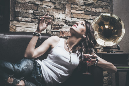 woman with glass of red wine and cigarette, wearing blue jeans and white t-shirt,  lie on leather couch, behin her is old gramophone, indoor shotの写真素材