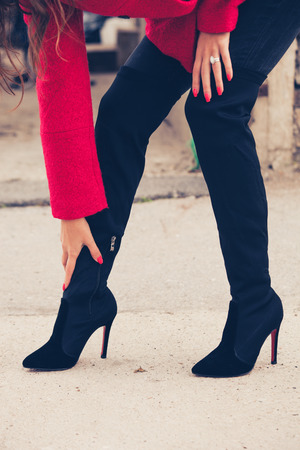 young woman on street in red coat touch her black high heels boots bent forward, closeup, selective focusの写真素材