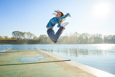 happy young woman jumping high  on pontoon at lake in  tracksuit, sunny autumn day, full body shotの写真素材