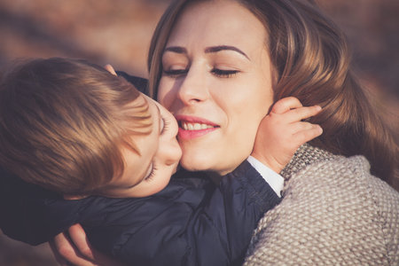 his mom is his greatest love, son and mom kissing and hugging, autumn day in park, closeup,...