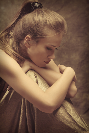 beautiful young sensual woman in golden dress sit with wrap  arms around legs, side view, studio shotの写真素材