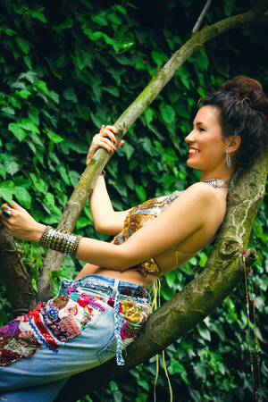 smiling young woman with bun sit on tree branch in boho style clothes blue embroidery jeans and golden topの写真素材