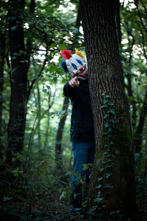 man with mask of scary clown stand behind tree in wood point with fingerの写真素材