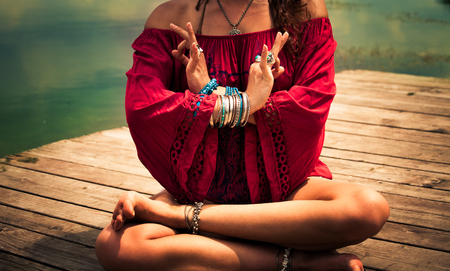 closeup young woman in a meditative yoga position wearing red tunic sit on wooden pontoon on the lake wearing lot of bracelets and ringsの写真素材