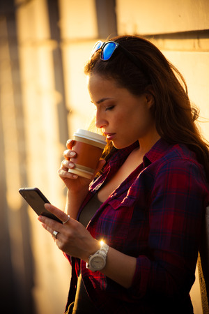 urban girl in city with smartphone and takeaway coffee lean on wall summer dayの写真素材