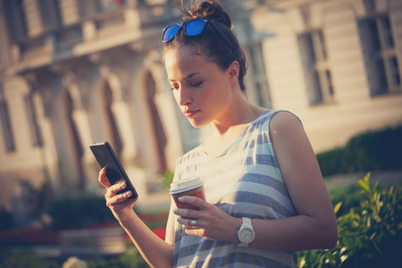 student girl in the city  streert using smartphone and drinking coffee wearin casual summer clothes and sunglasses on headの写真素材