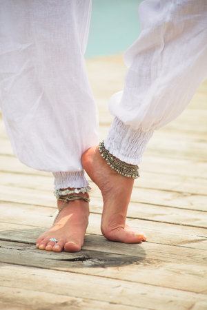 closeup of young woman barefoot legs in white clothes and anklets boho summer fashion style on...