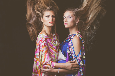 two blonde young women in embrace beauty fashion portrait with hair in motion in colorful silky dressの写真素材