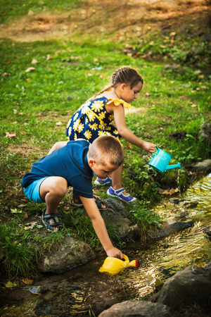 little boy and girl play draw water in bucket from stream outdoor summer day in natureの写真素材
