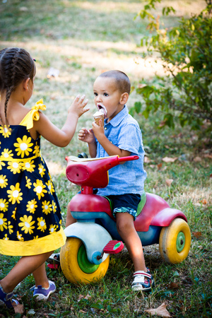 little boy eating ice cream sit on small tricycle while his sister approache him summer day in parkの写真素材